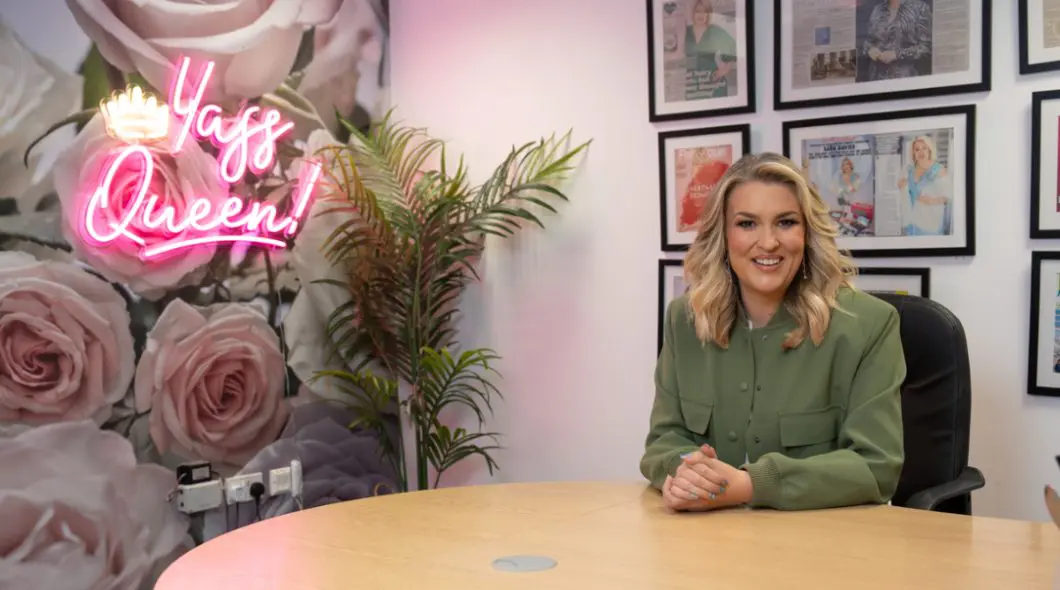 Sara Davies sat at a desk with a neon sign behind