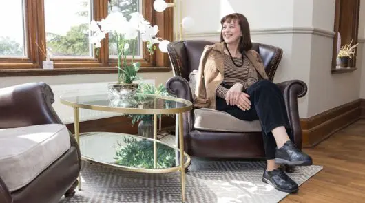 A woman in a brown jacket, top and black trousers sat in an armchair